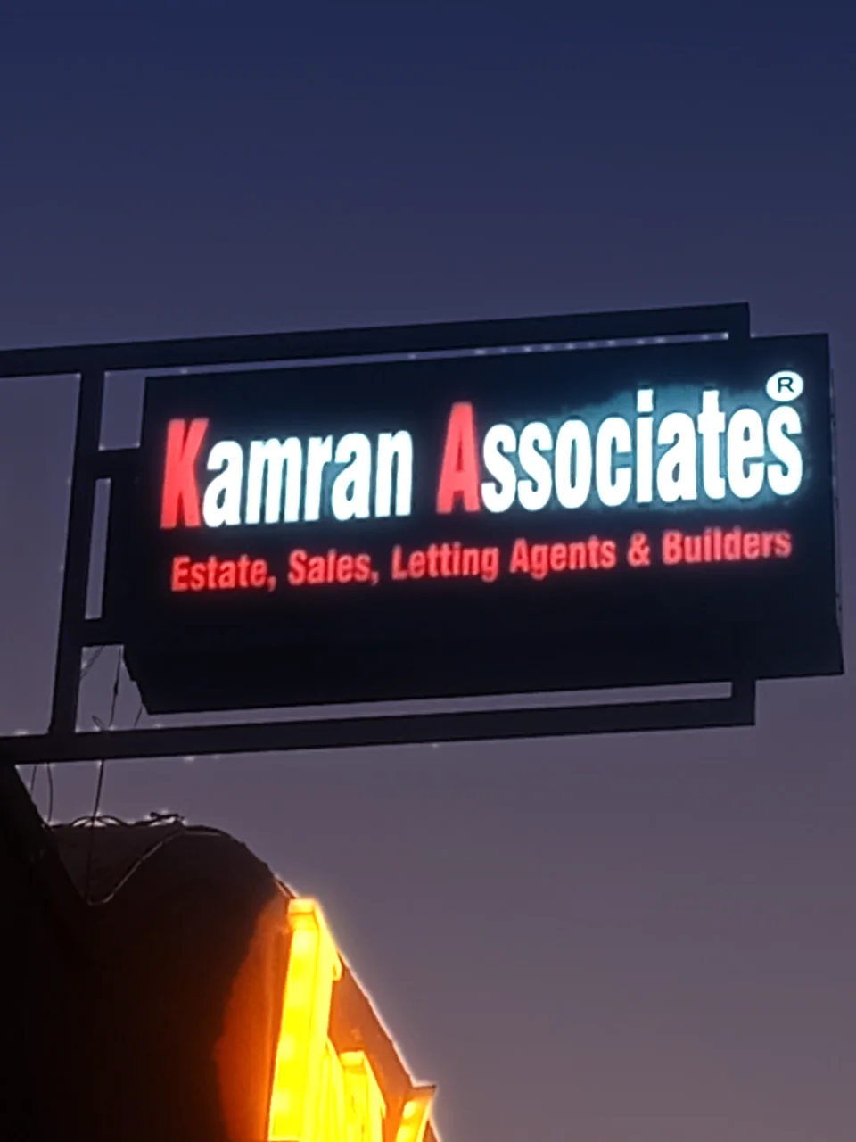 The Kamran Associates sign on the building at 99-B3 MM Alam Road, Gulberg III, lit at dusk